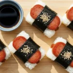 Homemade spam musubi wrapped in nori seaweed served on a wooden board with soy sauce dipping.